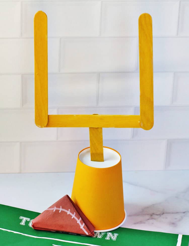 Football goal posts made from yellow craft sticks and a cup next to a paper folded to look like a football.