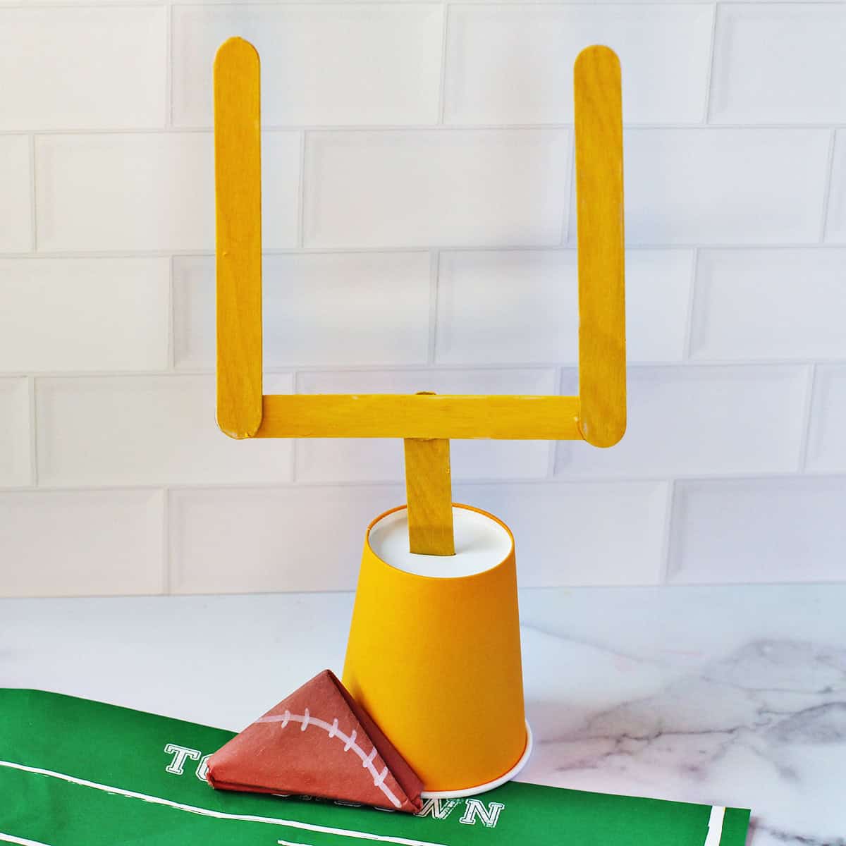 Football goal posts made from yellow craft sticks and a cup next to a paper folded to look like a football.