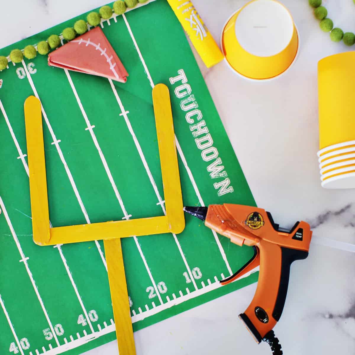 Hot glue gun sitting next to yellow craft sticks to look like a field goal post.