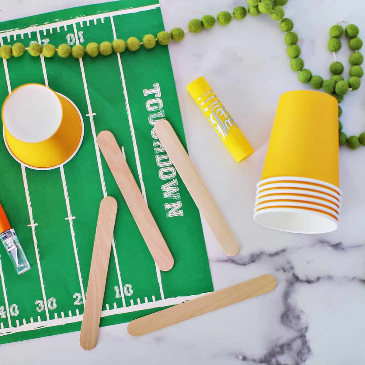 Yellow paper cup, craft sticks, an x-acto knife and a paint marker sitting on a placemat that looks like a football field.
