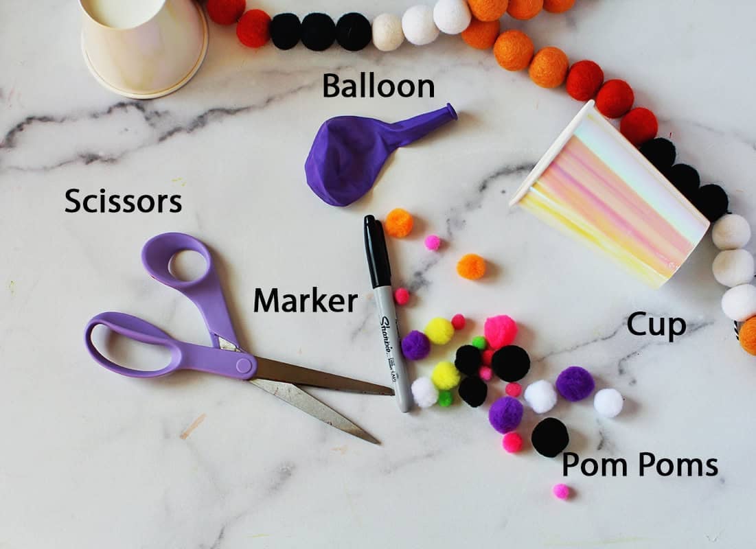 Purple scissors, purple balloon, black marker, pom poms and an iridescent cup on a counter.