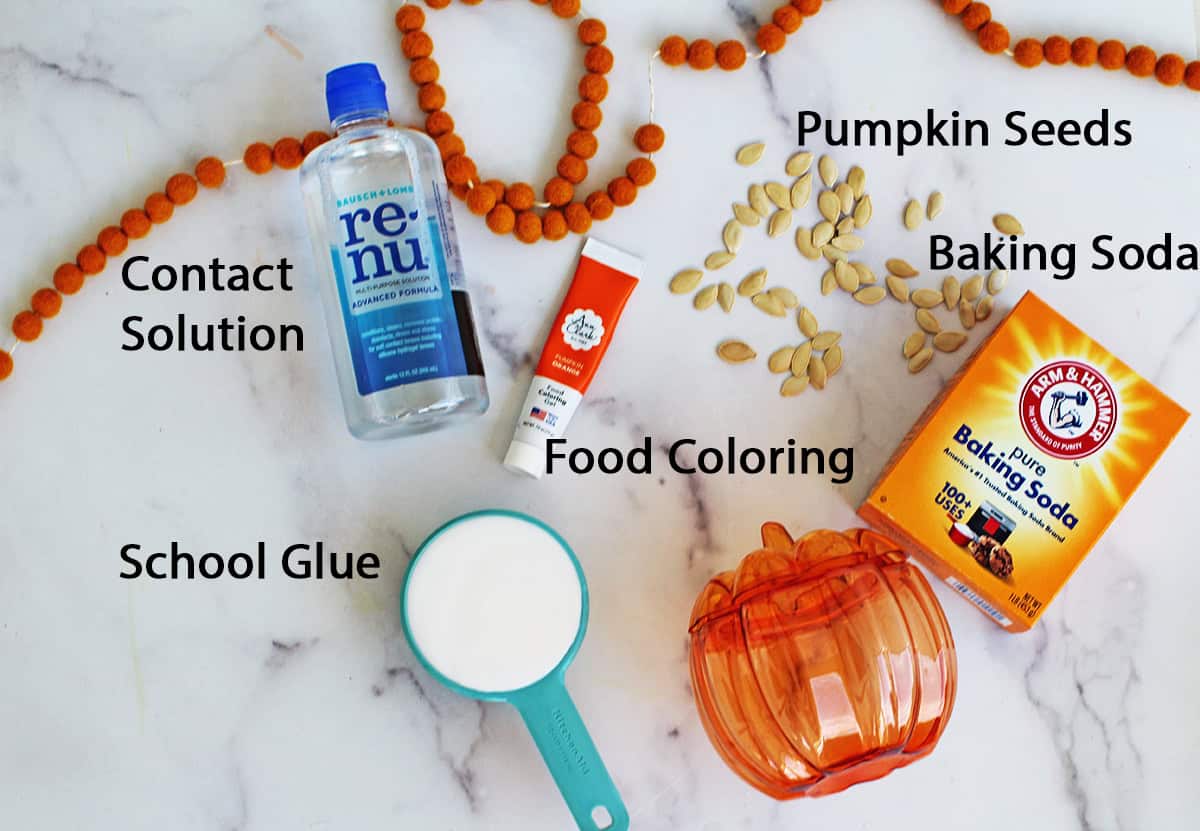 Baking soda, plastic pumpkin, school glue in a measuring cup, contact solution, pumpkin seeds and food coloring on a counter.