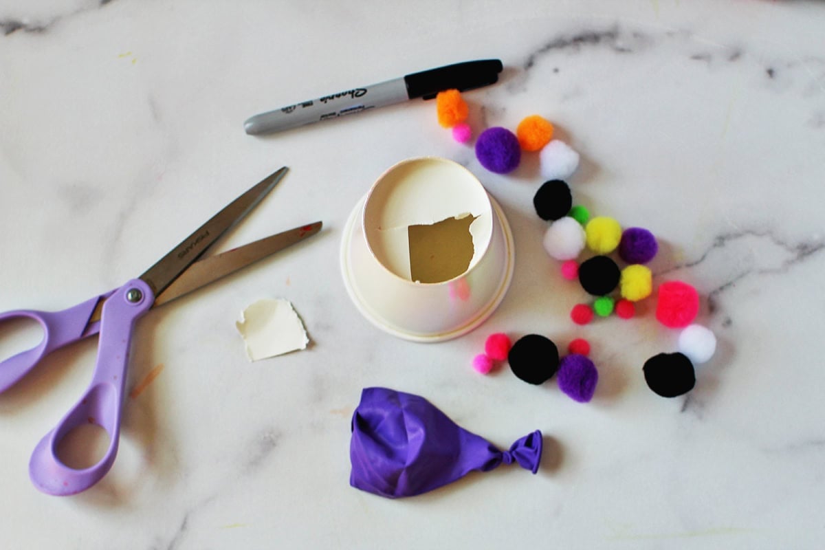 Cup that is having the bottom cut out next to a cut balloon and pom poms on a counter.