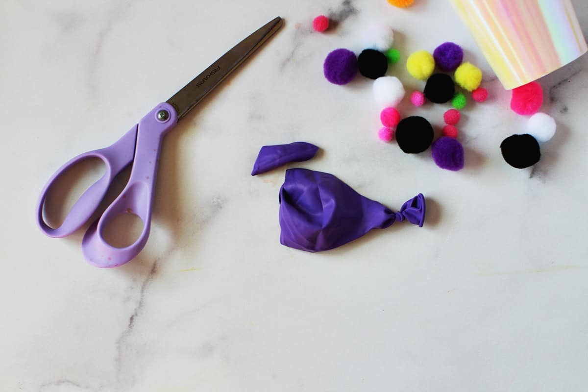 Purple scissors cutting the top off a tied purple balloon next to pom poms.
