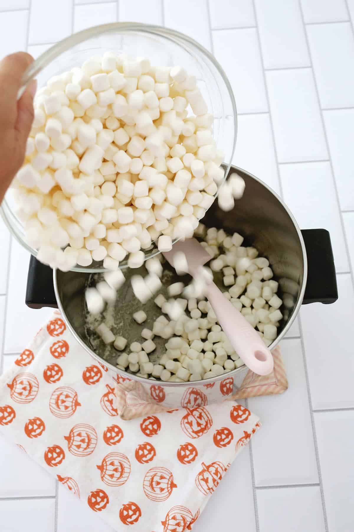 A bowl of mini marshmallows being poured into a pot to melt with butter and marshmallows for pumpkin shaped rice krispie treats.