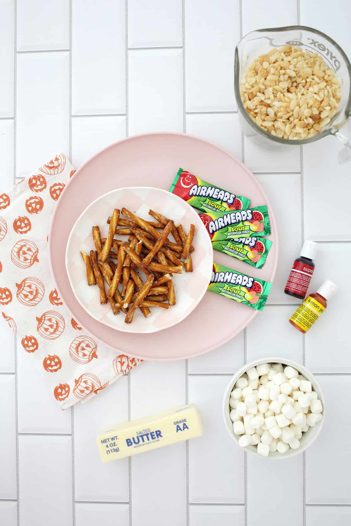 a pink plate with green airheads next ot a plate of pretzel rods on a counter. Also on the counter is a stick of butter, a bowl of mini marshmallows and food coloring to make rice krispies pumpkin shaped treats.