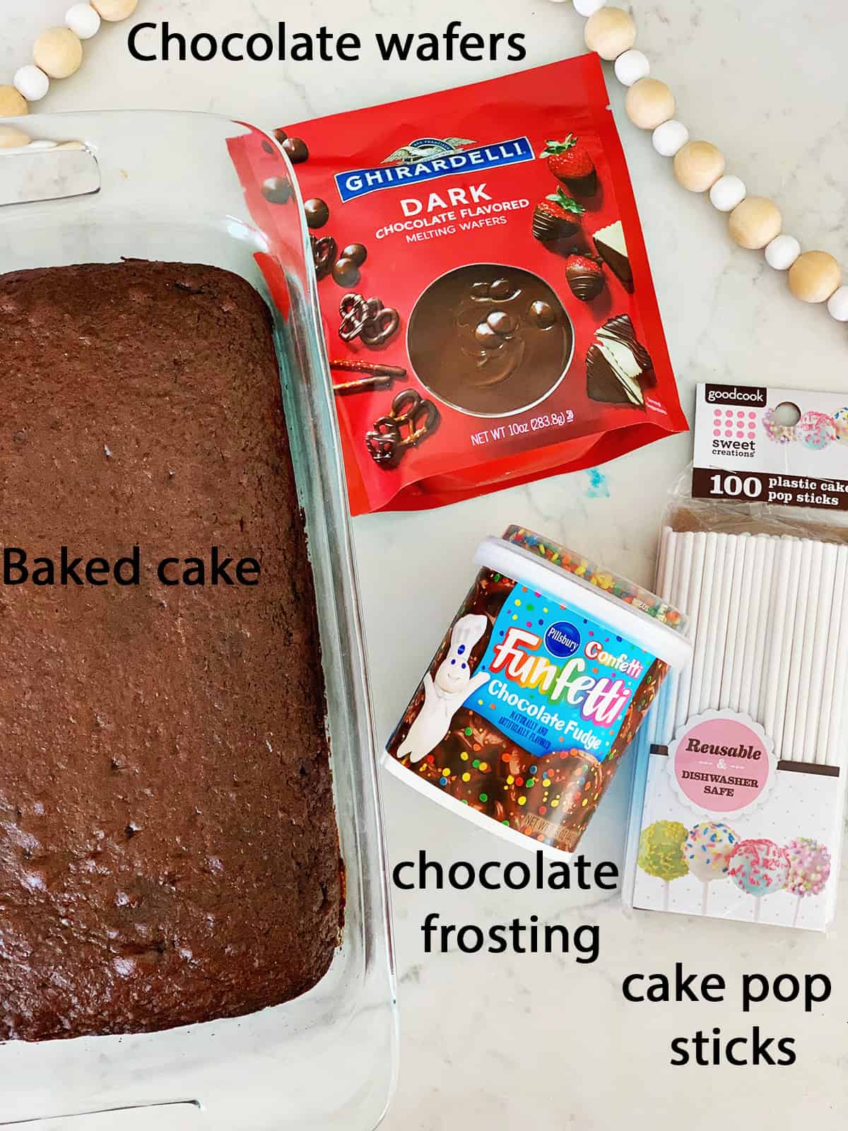 Chocolate cake next to cake pop sticks, chocolate frosting and chocolate melting wafers on a counter.