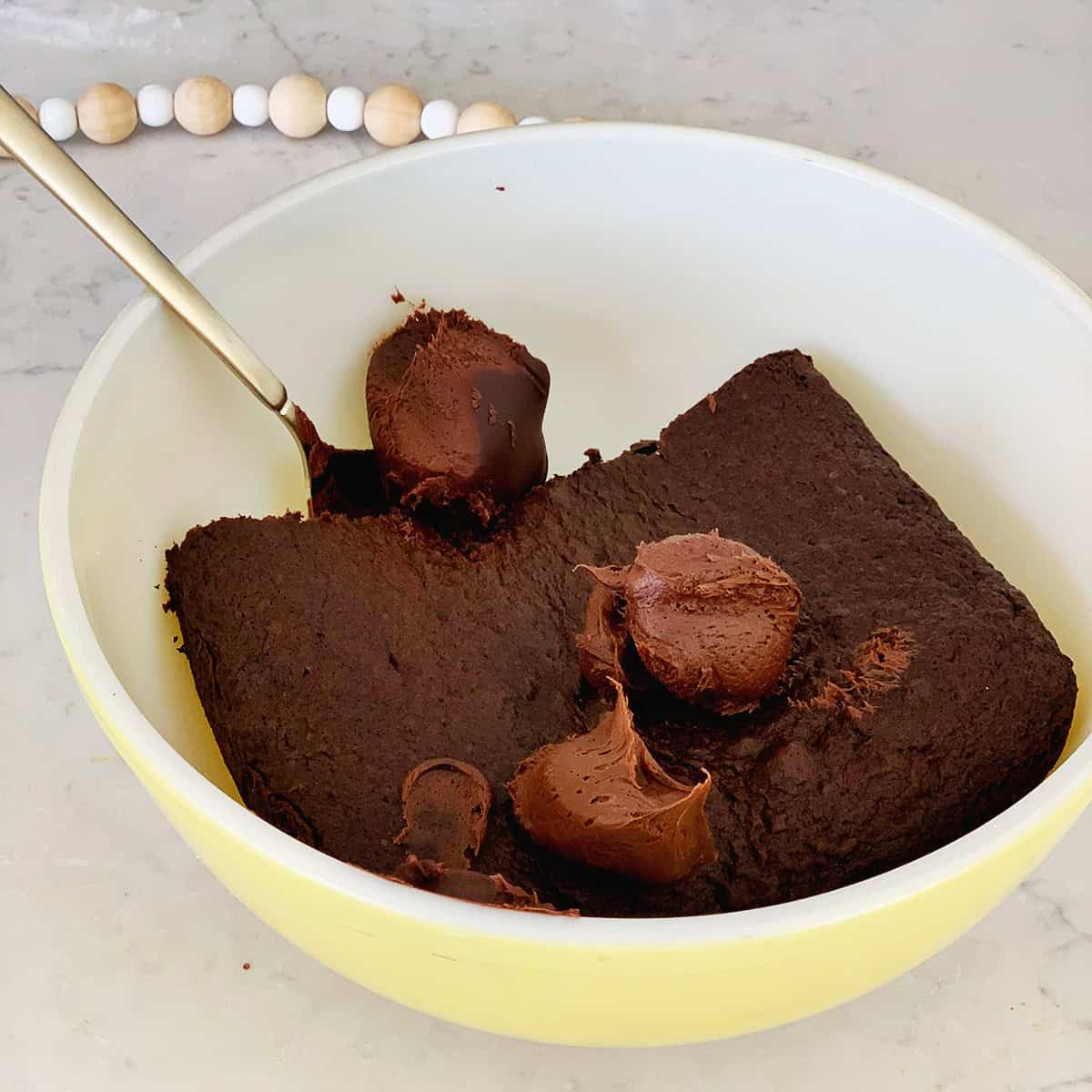chocolate frosting being added to a mixing bowl with a chocolate cake in it.